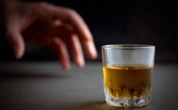 hand reaches for a glass of whiskey or cognac or alcohol drink, alcoholism and alcohol abuse concept, defocused, selective focus, close up, gray table, dark background